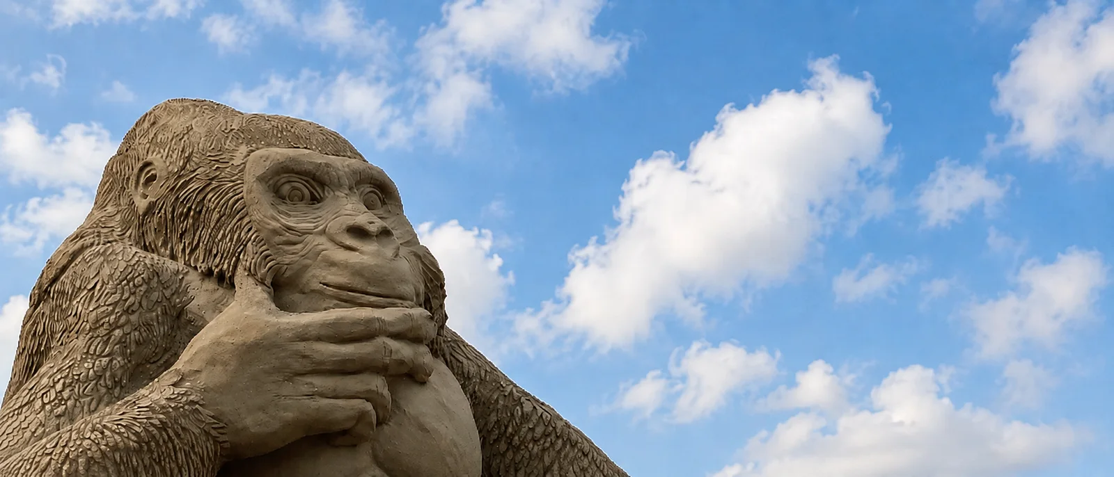 A sand sculpture of a great ape sitting in thought, gazing up at a bright blue, cloud-scattered sky